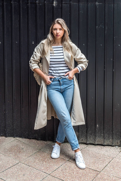 Portrait of attractive lady in checkered blue jeans and beige trench coat looks and poses outside. Fashionable woman outdoor posing in cozy stylish outfit  - Valokuva, kuva