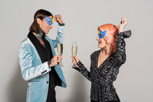 stylish and happy queer people in party masks dancing with champagne glasses on grey background - Photo, Image