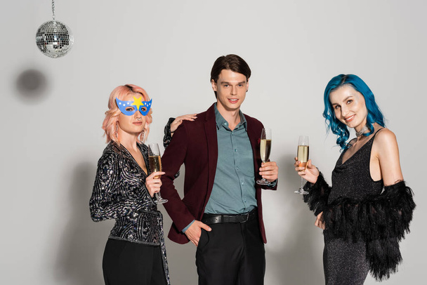 элегантные голубые друзья с бокалами для шампанского, улыбающиеся в камеру рядом с небинарным человеком, стоящим с рукой в кармане на сером фоне - Фото, изображение