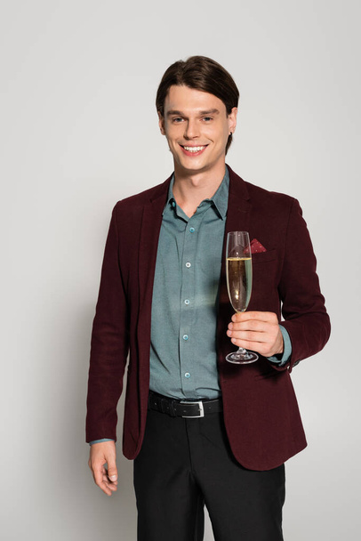 happy young man in brown blazer holding glass of champagne isolated on grey - Fotó, kép