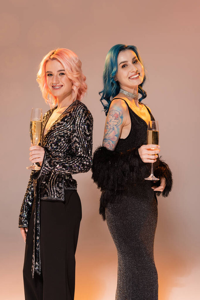 queer friends in festive elegant clothes holding champagne and smiling at camera on beige background - Photo, Image