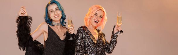 cheerful and elegant nonbinary friends with champagne glasses looking at camera on beige background, banner - Photo, Image