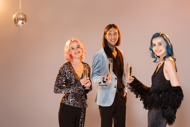 stylish and happy nonbinary people with champagne glasses near shiny disco ball on beige background - Photo, Image
