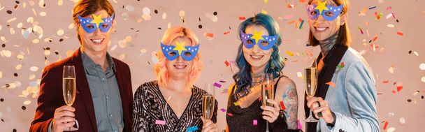 sonrientes amigos queer en máscaras de fiesta sosteniendo copas de champán bajo la caída de confeti sobre fondo beige, bandera - Foto, imagen