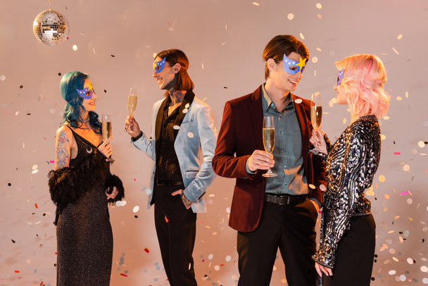 happy queer people in party masks holding champagne glasses and talking under falling confetti on beige background - Photo, Image