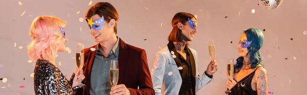 cheerful nonbinary people talking while standing with champagne glasses under falling confetti on beige background, banner - 写真・画像