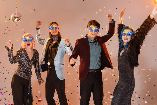 возбужденные голубые друзья в праздничной одежде и масках, танцующие во время новогодней вечеринки на бежевом фоне - Фото, изображение