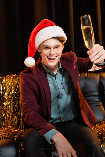 gelukkig man in santa hoed toosten met champagne terwijl zitten in de buurt gouden knutsel op donkere achtergrond - Foto, afbeelding