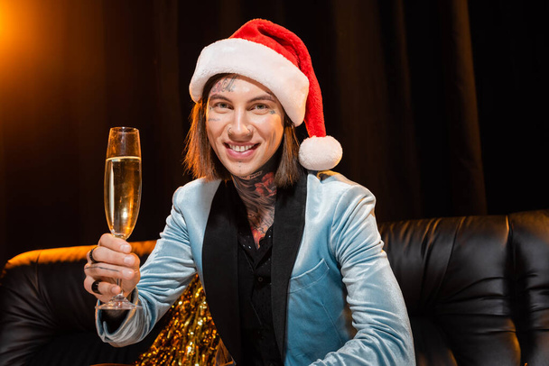 Tätowierte nichtbinäre Person mit Weihnachtsmütze, Champagner in der Hand und Blick in die Kamera auf dunklem Hintergrund mit Beleuchtung - Foto, Bild