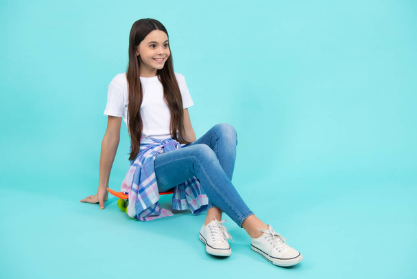 Teenager životní styl. Módní sebevědomá dívka v módním tričku a džínách sedí na skateboardu, pózuje na modrém pozadí. Módní teenager. Šťastný teenager, pozitivní a usměvavé emoce - Fotografie, Obrázek