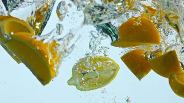 Φρέσκια νόστιμη πορτοκαλί σφήνες πέσει σε διαφανές νερό closeup. Κομμάτια φωτεινά ζουμερά εσπεριδοειδή επιπλέουν βαθιά κάτω από διαυγές υγρό με φυσαλίδες αέρα σε εξαιρετικά αργή κίνηση. Ωριμάζουν κίτρινα φρούτα για νόστιμο ποτό - Φωτογραφία, εικόνα