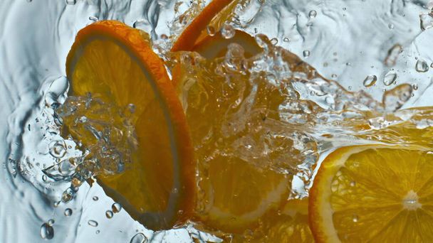 Kousky oranžové kapala čistá voda v super zpomaleném filmu zblízka. Plátky šťavnaté citrusové plody cákající v průhledné bublinkové vodě na bílém pozadí. Chutná vitamínová přísada pro osvěžující šťávu. - Fotografie, Obrázek