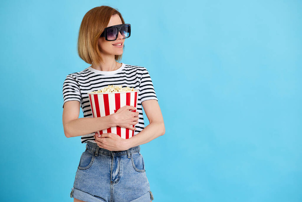 Wesoła młoda kobieta ogląda film, trzymając wiadro popcornu na niebieskim tle. przestrzeń kopiowania - Zdjęcie, obraz