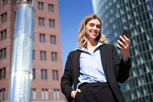 Niederwinkelaufnahme einer Geschäftsfrau im Anzug, die auf der Straße steht und auf ihr Handy schaut und ihr Smartphone in der Hand hält. - Foto, Bild