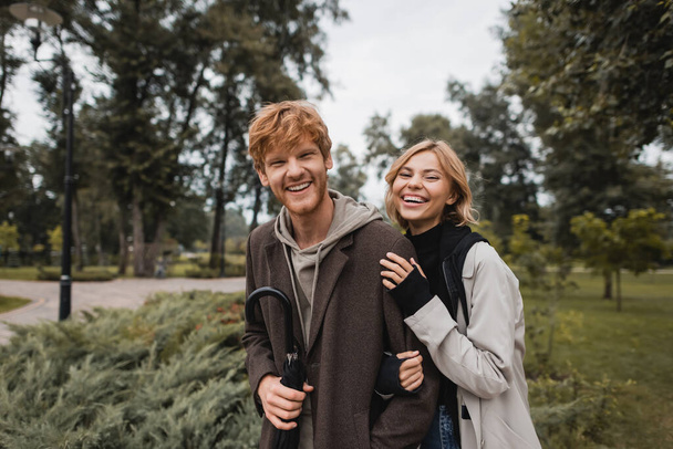 happy blonde woman hugging cheerful redhead boyfriend with umbrella  - Valokuva, kuva