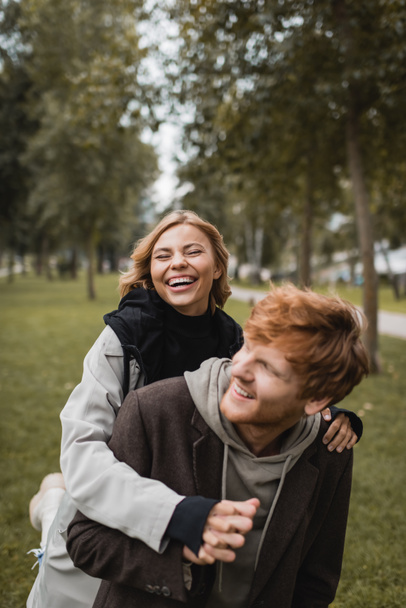 θετική νεαρή γυναίκα στο παλτό αγκαλιάζει χαρούμενο κοκκινομάλλα φίλο και γελά στο φθινοπωρινό πάρκο - Φωτογραφία, εικόνα