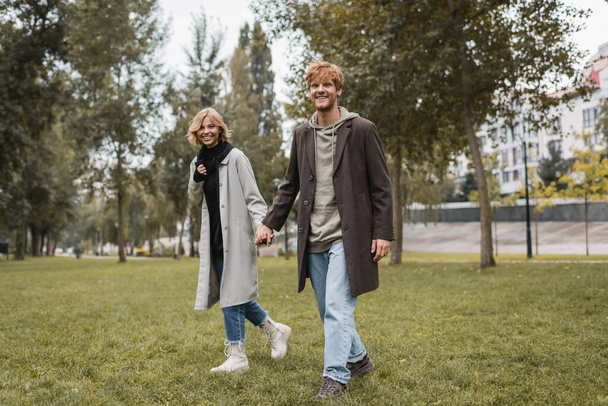 full length of happy young man in coat holding hands with cheerful girlfriend while walking in autumnal park  - Foto, afbeelding