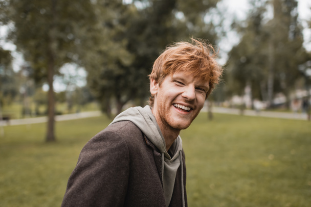 cheerful and redhead man in autumnal outfit smiling in park  - Fotó, kép