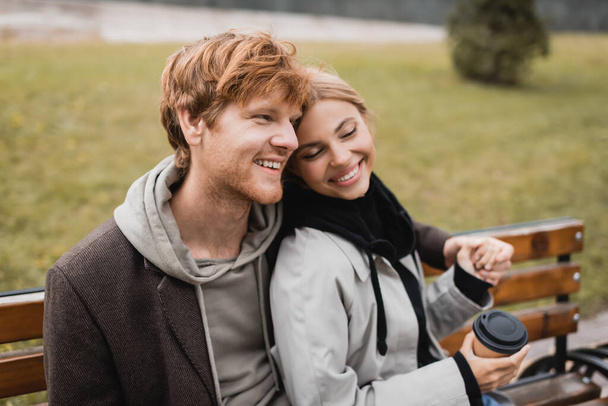 glücklicher junger Mann umarmt fröhliche Freundin mit Pappbecher, während er auf Bank sitzt - Foto, Bild