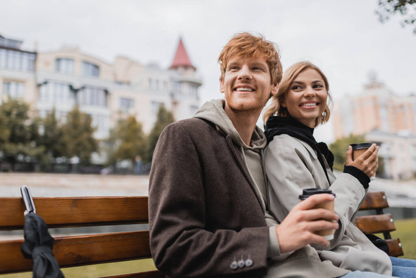 fröhliches junges Paar umarmt und hält Pappbecher mit Coffee to go in der Hand, während es auf einer Bank im Park sitzt - Foto, Bild