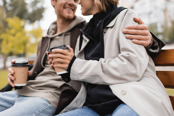 vue recadrée de heureux jeune couple étreignant et tenant des tasses en papier avec du café pour aller dans le parc - Photo, image