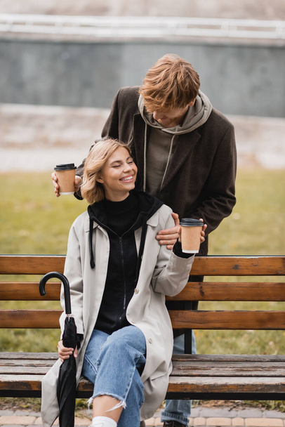 zrzka muž drží papírové poháry v blízkosti blondýny žena sedí na lavičce s deštníkem v parku - Fotografie, Obrázek