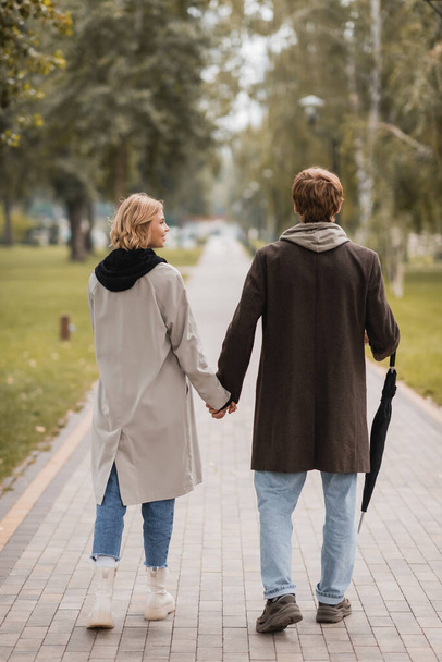 Rückansicht von Paar in Mänteln, Händchen haltend und im herbstlichen Park spazierend  - Foto, Bild