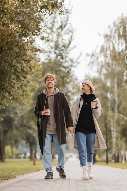full length of happy couple in coats holding hands and walking with paper cups in autumnal park  - Photo, image