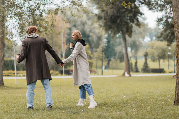 full length of redhead young man in coat holding paper cup and hand of happy girlfriend in autumnal park  - Foto, imagen