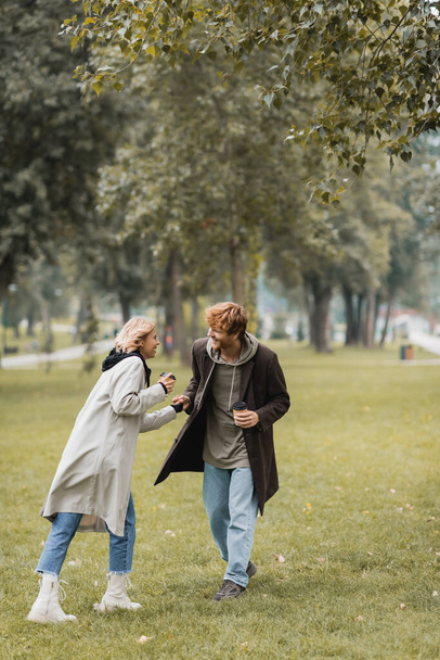 comprimento total de homem alegre no casaco segurando copo de papel e mão da namorada satisfeita no parque outonal  - Foto, Imagem