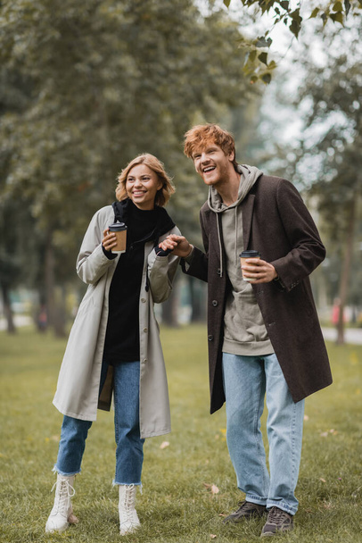 full length of positive man with paper cup holding hands with pleased girlfriend in autumnal park  - Foto, Bild