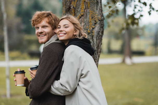 cheerful blonde woman holding paper cup and hugging with boyfriend near tree trunk - Photo, Image