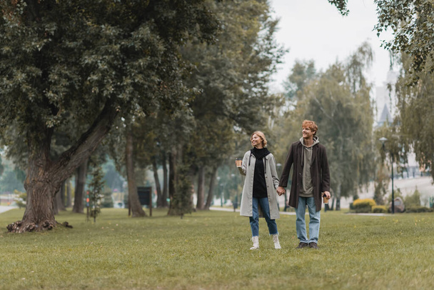 pełna długość szczęśliwy ruda mężczyzna i wesoły kobieta trzymając kawę iść podczas spaceru w parku - Zdjęcie, obraz