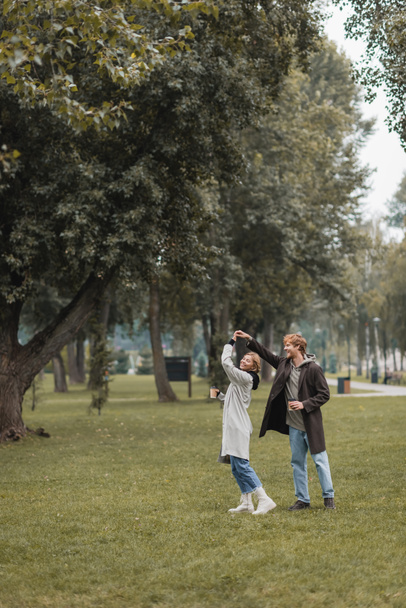 pleine longueur de rousse heureux homme et femme gaie tenant café pour aller tout en dansant dans le parc - Photo, image