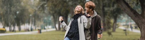 happy redhead man looking at cheerful woman holding coffee to go while laughing in park, banner - 写真・画像