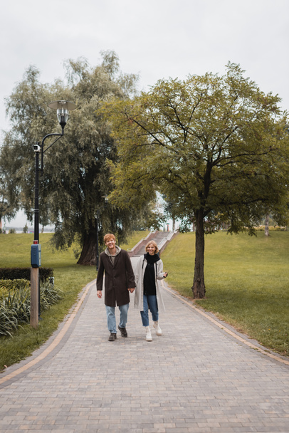 volledige lengte van vrolijke roodharige man en tevreden jonge vrouw in jas hand vasthouden tijdens het wandelen in het park  - Foto, afbeelding