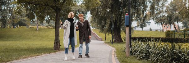 full length of happy redhead man and blonde young woman in coat holding hands while walking in park, banner - Foto, afbeelding