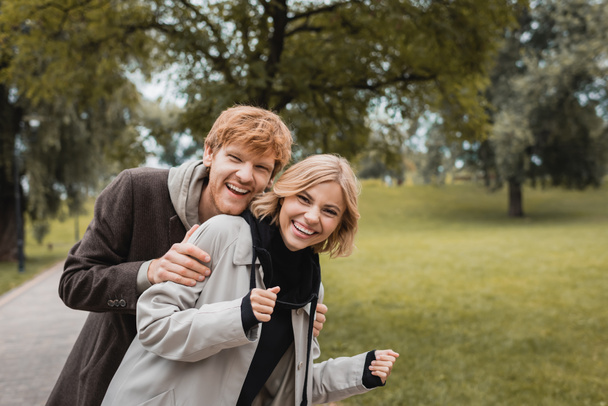 happy man hugging joyful young woman while looking at camera in park  - Photo, Image