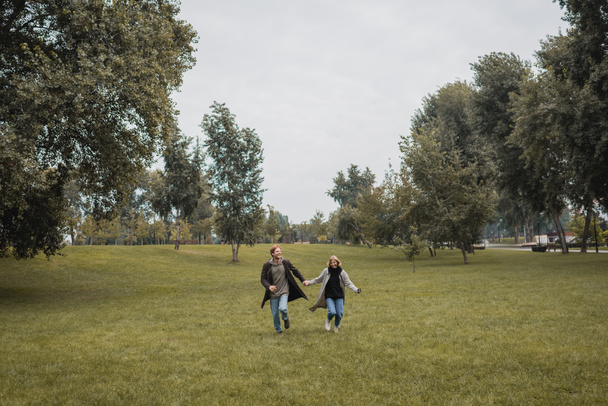на всю длину счастливого мужчины и довольной женщины в пальто, держась за руки во время бега по траве  - Фото, изображение