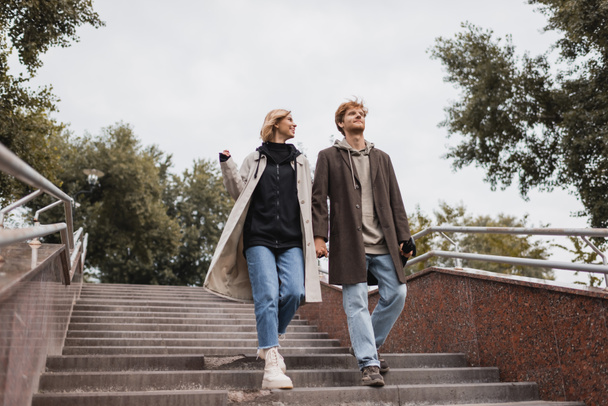 low angle view of cheerful woman and redhead man with umbrella holding hands while descending stairs near park - Foto, afbeelding