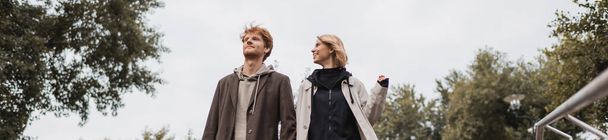 low angle view of cheerful woman and redhead man against cloudy sky in park, banner - Foto, imagen