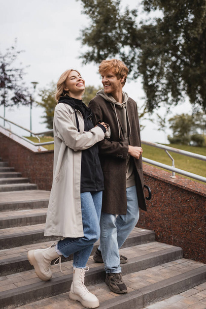 joyful woman and happy redhead man with umbrella descending stairs in park - 写真・画像