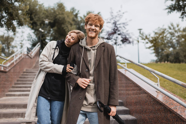 pleased woman leaning on shoulder of redhead man with umbrella while descending stairs in park - Photo, Image