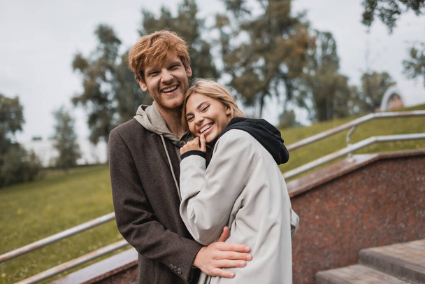 happy young woman hugging with joyful redhead boyfriend in autumnal coat in park - Photo, Image