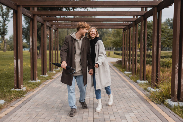 full length of happy young couple in autumnal coats holding hands while walking under multiple arch in park - Photo, Image