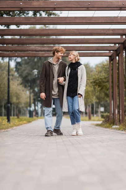 πλήρες μήκος του ευτυχισμένου ζευγαριού σε φθινοπωρινά παλτά κρατώντας τα χέρια, ενώ το περπάτημα κάτω από πολλαπλές αψίδα στο πάρκο - Φωτογραφία, εικόνα