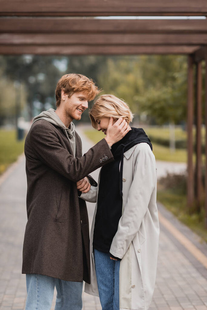 happy young man in autumnal coat hugging woman under arch in park - Photo, Image