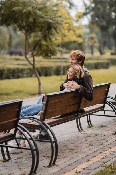 Neşeli genç bir adam kadına sarılıyor ve parktaki ahşap bankta oturuyor. - Fotoğraf, Görsel