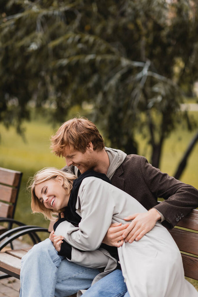 redhead man laughing while hugging happy girlfriend while sitting on bench - Fotografie, Obrázek