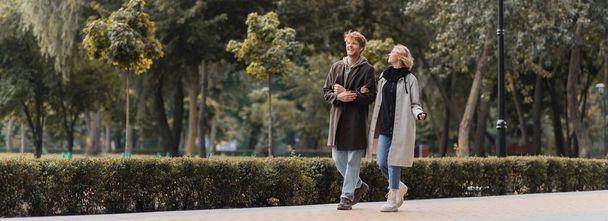 full length of redhead man and blonde woman in coat smiling while walking in park, banner - Photo, image